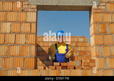 Mason tragen gelbes T-shirt und blaue Helm mit Edelstahl-Kelle posiert unter unfertige Haus Ziegelwände Stockfoto