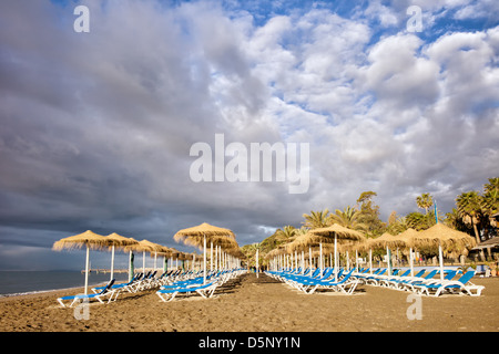 Liegestühle am Morgen an einem Sandstrand am Mittelmeer im beliebten Ferienort Marbella in Spanien. Stockfoto