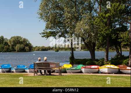 Ein paar sitzt auf eine hölzerne Bank mit Blick auf Thorpeness Meare - eine traditionelle Bootsteich in Suffolk, England, UK Stockfoto