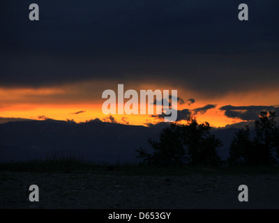 Ein dramatischer Sonnenuntergang über bergigem Gelände mit Sturmwolken am Himmel. Die leuchtenden Farben der untergehenden Sonne stehen im Kontrast zu den dunklen Wolken und schaffen eine kraftvolle natürliche Szene. Stockfoto
