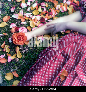 eine Mädchen in einem roten Kleid ist auf dem Rasen zwischen Blütenblätter, eine Rose in der Hand liegen. Stockfoto