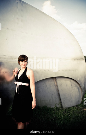 Junge, lächelnde Frau in schwarz Halfter Kleid mit rosa Schleife gelehnt Metallskulptur Stockfoto