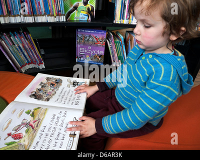 Ein 3 Jahre alter Junge Kind Lesen eines Tony Ross Bild Buch sitzend durch ein Regal in der Bibliothek Leseecke in Wales UK KATHY DEWITT