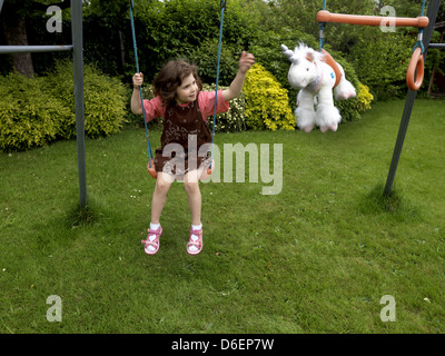 6 Jahre altes Mädchen auf der Schaukel im Garten mit ihrem Einhorn Plüschtier auf den Reifen neben ihr England Stockfoto