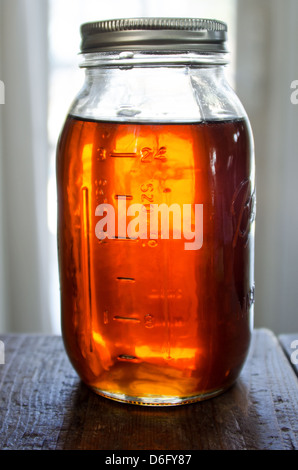 Ein Glas voll von Maine Ahornsirup Maurer steht auf einem rustikalen Holztisch vor einem sonnigen Fenster. Stockfoto