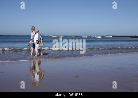 Ein älteres Ehepaar Fuß in den Wellen am Strand in Cadiz, Spanien.  Angelboote/Fischerboote auf das Atlantische Meer hinter schweben. Stockfoto