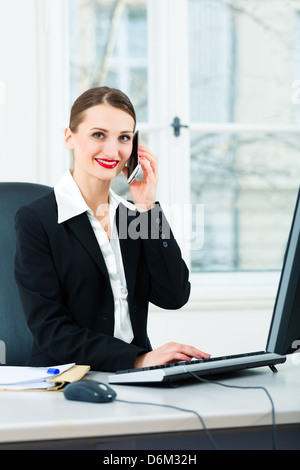 Junge Geschäftsfrau in ihrem Büro, sie sitzt auf dem Schreibtisch und am Telefon ist, ein Kunde oder Klient Stockfoto