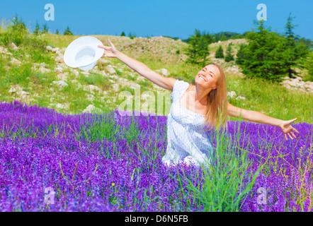 Hübsche Mädchen sitzen auf lila Lavendelfeld, warme Sonnenstrahlen genießen, halten in der hand weiße Sommerhut, Sommer-Natur Stockfoto