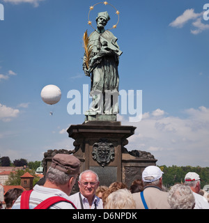 Touristen drängen sich unter die Staue des Hl. Johannes auf der Karlsbrücke, Prag. Ein Heißluftballon fliegt über den Fluss. Stockfoto