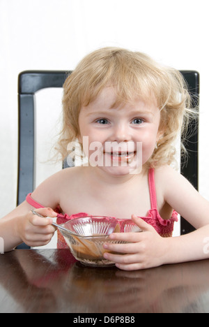 Junges Mädchen am Tisch einen Schokoladenpudding Essen und Lachen im Gesicht chaotisch: Studioportrait isoliert auf weiss Stockfoto