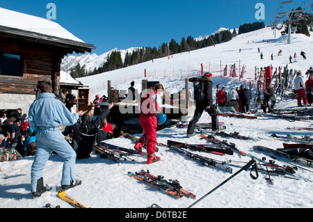 Familien halten zum Mittagessen in einem Bergrestaurant im Wintersportort La Clusaz in den französischen Alpen an, während Skifahrer im Hintergrund den Berg hinunterfahren Stockfoto