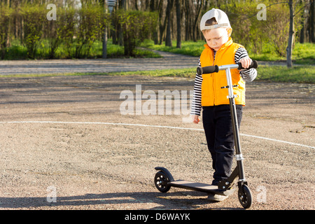 Hübscher kleiner Junge mit seinem Roller stehen auf einer geteerten Landstrasse blickte auf seine Füße mit einem gereizte Ausdruck in den Abend Sonnenlicht. Stockfoto