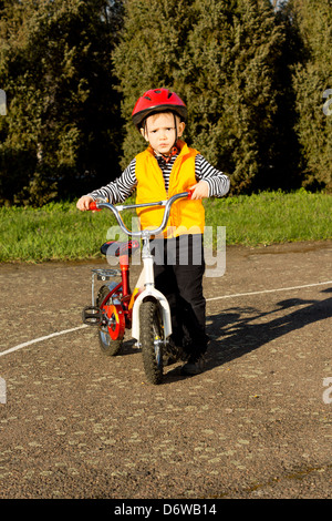 Entzückender junge gekleidet in eine leuchtend rote Helm und Orange hohe Sichtbarkeit Sicherheitsweste posiert stolz mit seinem Fahrrad auf der Suche auf den Betrachter. Stockfoto