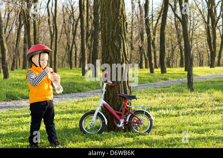 Kleiner Junge, mit seinem Fahrrad stoppen zwischen den Bäumen in einem bewaldeten Park für einen Schluck Wasser aus einer Plastikflasche Stockfoto