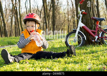 Junge Radfahrer genießen ein Glas erfrischendes Wasser aus einer Plastikflasche sitzen auf dem grünen Rasen unter den Bäumen in einem Naturpark mit seinem Fahrrad gelehnt auf einem Baum in der Nähe. Stockfoto