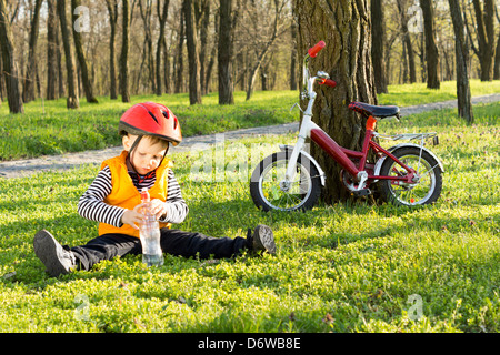 Süße junge auf seinem Fahrrad anhalten für einen Schluck Wasser in Flaschen sitzen auf saftig grünen Rasen in einem bewaldeten Park mit seinem Fahrrad gelehnt an einem Baum in der Nähe Reiten. Stockfoto