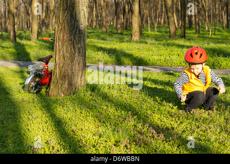 Kleiner Junge erkunden den Park kauerte auf dem grünen Rasen in einem Wäldchen, während sein Fahrrad lehnt sich gegen einen Baumstamm in der Nähe. Stockfoto