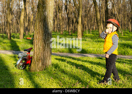 Kleiner Junge auf seinem Fahrrad erkunden gekleidet in einen Helm und Orange hohe Sichtbarkeit Sicherheitsweste in der Sonne zwischen den Bäumen in einem bewaldeten Park mit seinem Fahrrad an einen Baumstamm gelehnt. Stockfoto