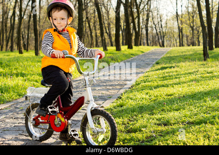 Entzückende kleine Junge mit seinem Fahrrad auf einem asphaltierten Weg in einen bewaldeten Park anhalten zu stehen und zu posieren für die Kamera in seine Sicherheit Stockfoto
