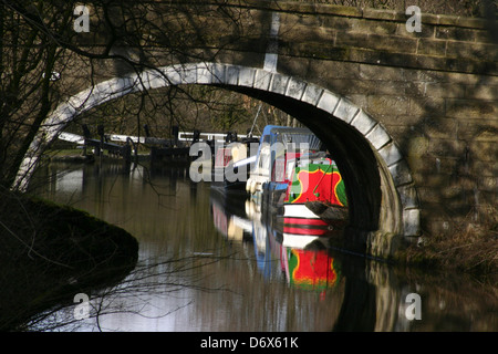 Ein rotes Kanalboot, das durch eine Steinbrücke auf dem Kanal Leeds und Liverpool mit Schleusen in der Ferne an der Bank Newton in North Yorkshire gesehen wird. Stockfoto