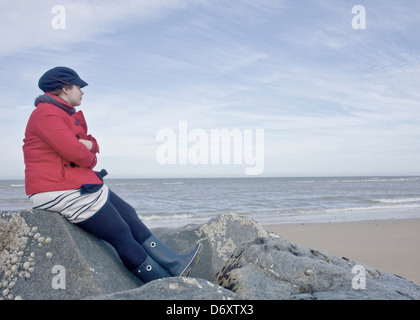 Frau im roten Mantel, sitzen auf den Felsen am Strand, Blick auf das Meer nachdenklich. Stockfoto