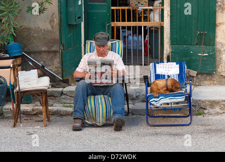 Saignon, Frankreich, ein älterer Mann sitzt mit seinem Hund am Straßenrand und liest Zeitung Stockfoto