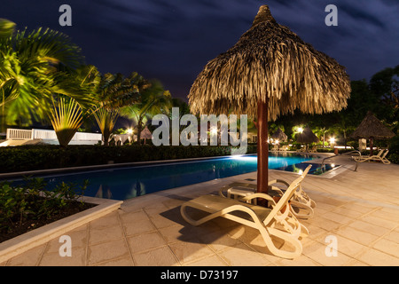 Strohschirm und zwei Beach-Plätze in der Nähe von einem beleuchteten Pool bei Nacht Stockfoto