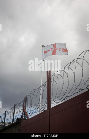 Mit Spulen Barde Sicherheit unter übersieht ein traurig aussehende englische Flagge an einem Mast einen industrielle Hof in Süd-London. Stockfoto