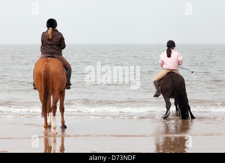 Zwei junge Frauen auf Pferden reiten am Strand von den Gewässern Rand Holkham Beach-Norfolk UK Stockfoto