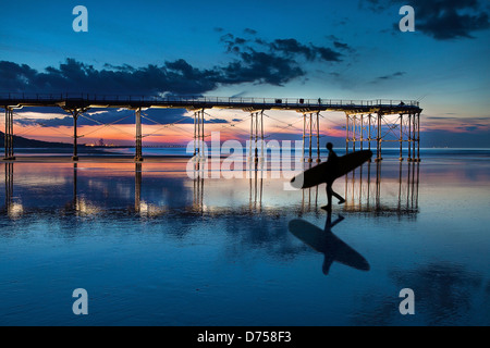 Saltburn Pier bei Sonnenuntergang als Surfer geht vorbei mit seinem Spiegelbild im nassen sand Stockfoto