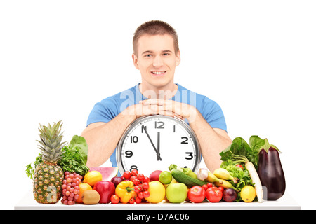 Hübscher Mann hält eine Wanduhr, sitzt auf einem Tisch voller Obst und Gemüse, isoliert auf weißem Hintergrund Stockfoto