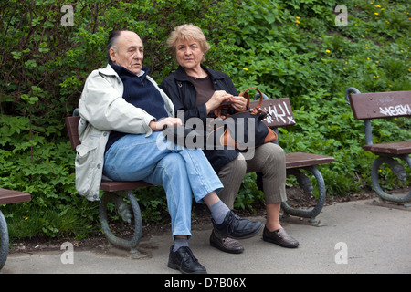 Senior Pärchen sitzen zusammen auf einer Parkbank im Freien, ruhen sich aus und unterhalten sich in einer grünen städtischen Umgebung Stockfoto