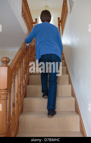 Alltägliche Szene einer einsamen älteren Frau, die Treppe hinauf geht und an einem hölzernen Geländer hält, der zu Hause von unten gesehen wird. England, Großbritannien Stockfoto