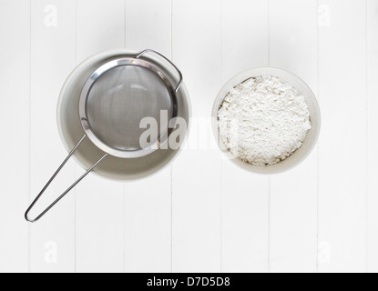 Eine Keramikschale mit weißem Mehl sitzt bereit, (gesiebt) Obwohl ein Metallsieb auf weißem Holz gesichtet werden. Stockfoto