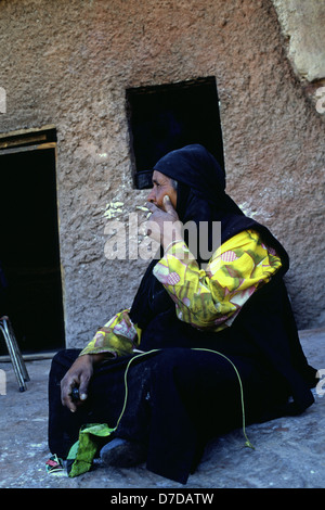 Eine ältere Frau aus der Beduinen Bedul Stamm einer Der Huwaitat Stämmen, die historisch in Petra gelebt haben, die traditionelle Kleidung das Rauchen einer Zigarette am Eingang zu einer Höhle Residence in Felsen gehauene von Wadi Musa in der Nähe von Petra Jordanien Stockfoto