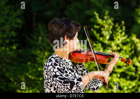 Horizontale Foto von Senior Asiatin zur Waldseite beim Spielen der Violine im Freien mit grünen Bäumen im Hintergrund Stockfoto