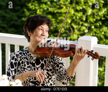 Horizontale Foto Senior asiatische Frau sitzen beim Spielen der Violine im Freien mit grünen Bäumen im Hintergrund Stockfoto