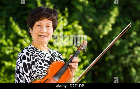 Horizontale Foto Senior asiatische Frau, die eine Pause vom Spielen der Violine im Freien mit leuchtend grünen Bäumen im Hintergrund Stockfoto