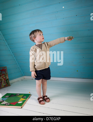 Ein kleiner Junge in schöne Kleider und Sandalen spielt mit alten altmodischen Brettspiel in einer blau-weißen Loft-Zimmer allein. Stockfoto