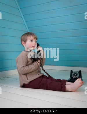 Ein Junge spielt mit einem alten altmodischen Wählscheibe Telefon sitzen auf dem weißen Holzboden barfuß in einem blauen Loft-Zimmer Stockfoto