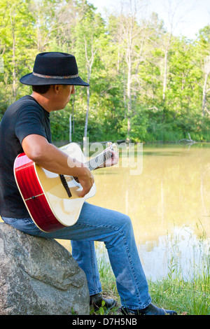 ein Mann in einem Cowboy-Hut spielt eine akustische Gitarre nach außen in einen ruhigen Teich in Sarnia, Ontario, Kanada Stockfoto