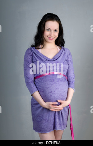 lächelnde junge Frau in ihren 20er Jahren, neun Monaten schwanger in lila Kleid auf grauem Hintergrund Modell veröffentlicht Stockfoto