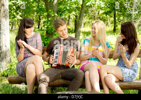 Glückliche junge Frau Freunde klatschten entlang zur Musik auf ein Akkordeon durch ein hübscher junger Mann gespielt, wie sie einen entspannten Tag im Park genießen. Stockfoto