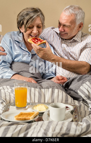 Älteres Paar im Bett Mann Fütterung Frühstück Frau liegend Stockfoto