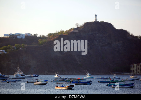 Die Abenddämmerung lässt sich über San Juan del Sur, Nicaragua, nieder, während Fischerboote im Hafen unter einer Statue auf einem Hügel ruhen. Stockfoto