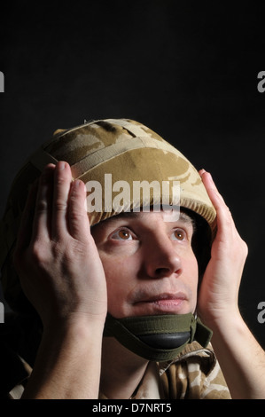 Soldat mit den Händen auf Helm vor einem dunklen Hintergrund. Soldat könnte von PTBS leiden. Stockfoto