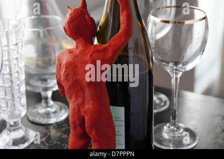 Temptatia kleiner roter Teufel, (Modelliermasse) von hinten gesehen, versteckt sich hinter einer Flasche Wein. Einige Gläser sind auf einem eingelegten Tisch verteilt Stockfoto