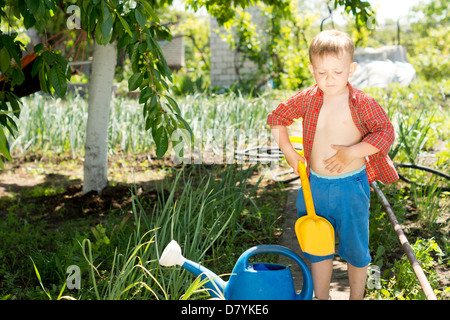 Kleiner Junge mit einem bunten gelben Plastikspielzeug Spaten im Garten stehen neben einer blauen Gießkanne unter das Gemüse Stockfoto