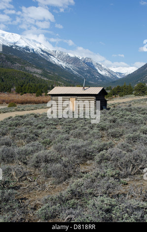 Eine alte verlassene Blockhütte befindet sich mit einem atemberaubenden Blick auf den schneebedeckten Rocky Mountains von Colorado. Stockfoto