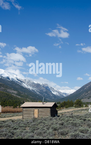 Eine alte verlassene Blockhütte befindet sich mit einem atemberaubenden Blick auf den schneebedeckten Rocky Mountains von Colorado. Stockfoto